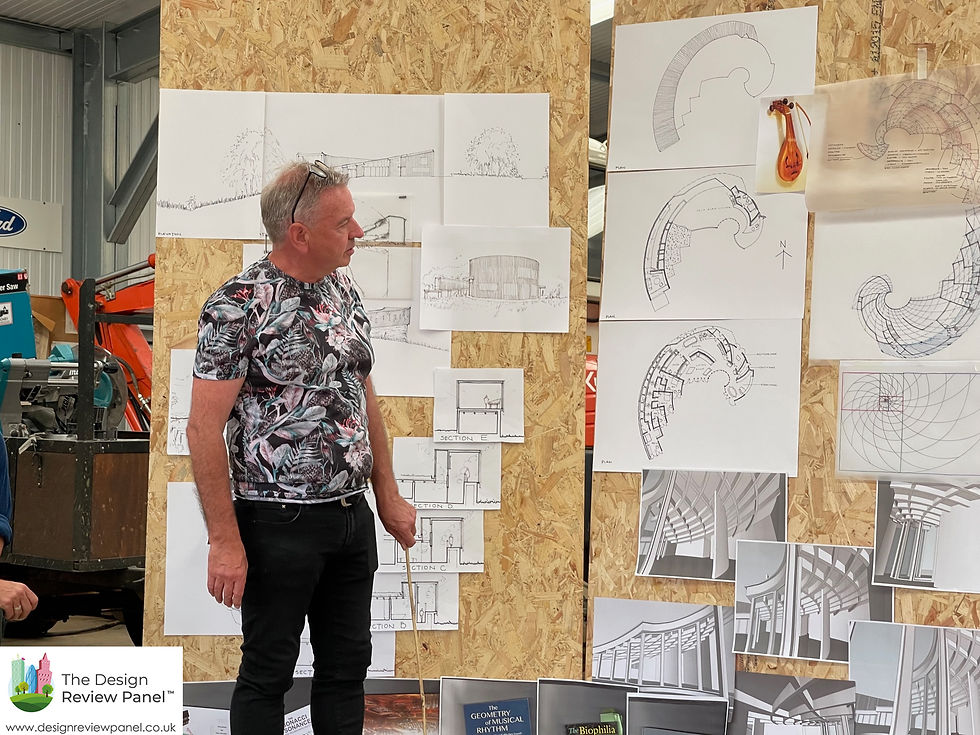 Man in floral shirt presents architectural sketches pinned on a corkboard in an industrial setting. Diagrams are detailed and complex.