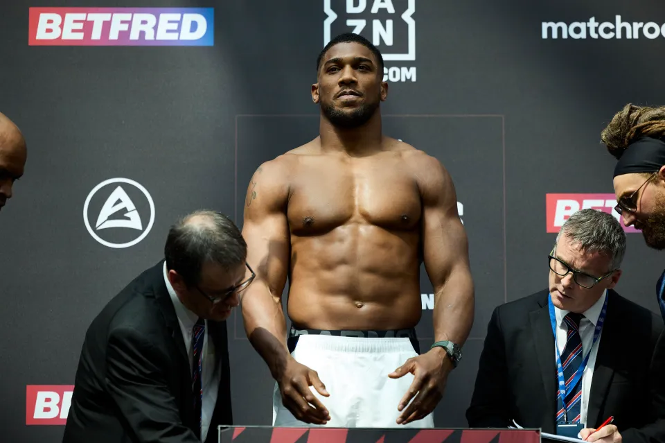 Shirtless man stands on a scale at an event, surrounded by officials. Bold black and red branding in background, focused atmosphere.