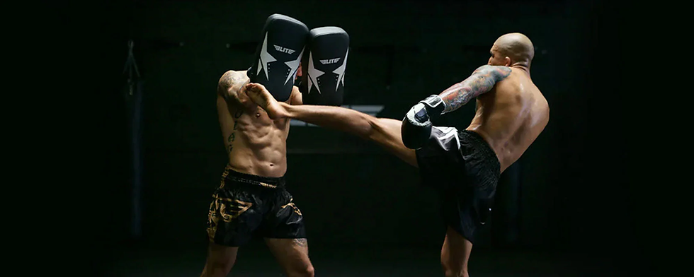 Two shirtless fighters in a dark gym; one throws a powerful kick, the other blocks with pads labeled "Elite.” Intense scene.