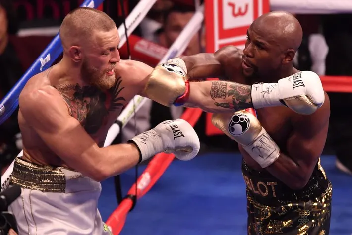 Two boxers exchange punches in a boxing ring. One wears white shorts and the other black. Emotions are intense, with a cheering crowd.