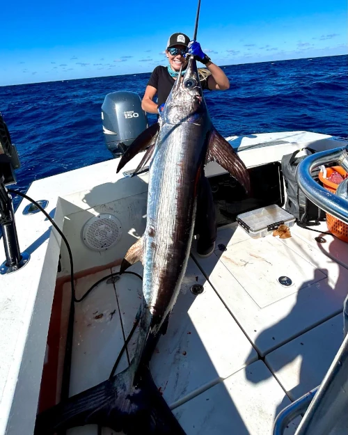 Person holding a large swordfish proudly on a boat, surrounded by ocean. Clear blue sky, calm sea, and boat equipment are visible.