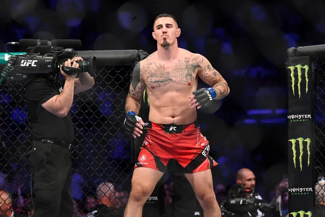 MMA fighter in red shorts and gloves stands confidently in a ring with a tattooed torso. A cameraman films nearby. Monster logo visible.