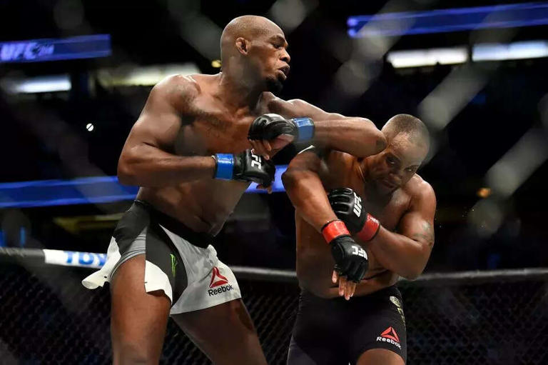 Two MMA fighters in a cage, one punching the other. Both wearing black gloves and shorts with Reebok logos. Intense action in the ring.