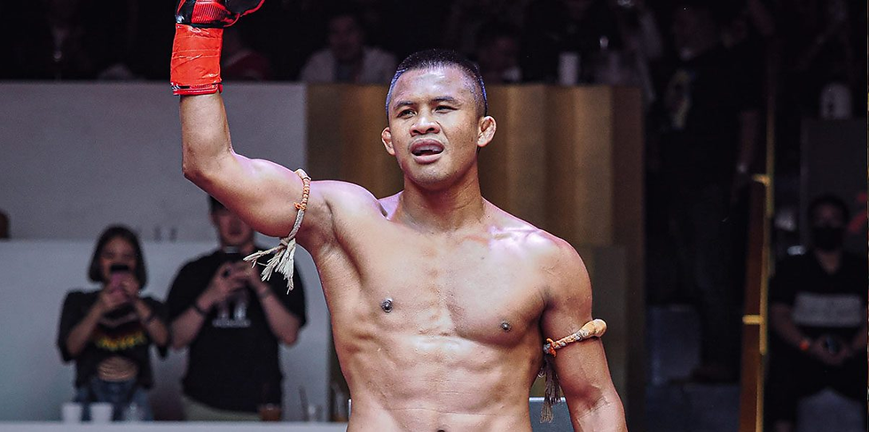 Shirtless man raises gloved fist in a victory pose, wearing armbands in a dimly lit indoor setting. Crowd in background. Confident expression.