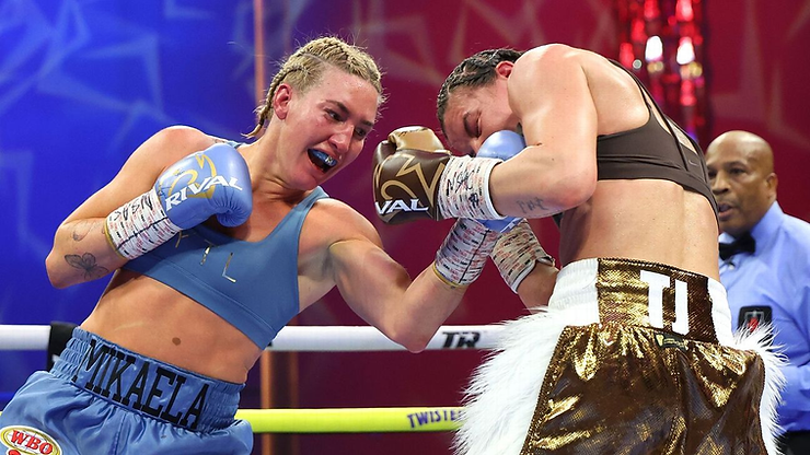 Boxer in blue punches opponent in brown during a match. Referee watches closely. Mikaela is visible on shorts. Intense action scene.