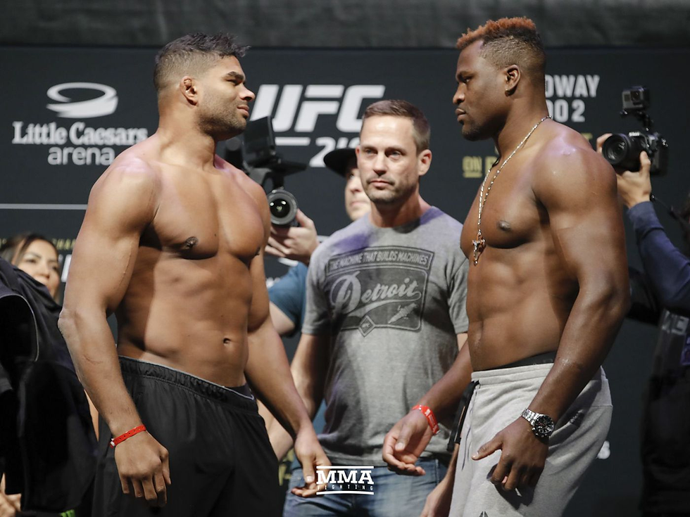 Two muscular men face off in a UFC weigh-in. One wears black shorts, the other gray. Background displays Little Caesars Arena and text.