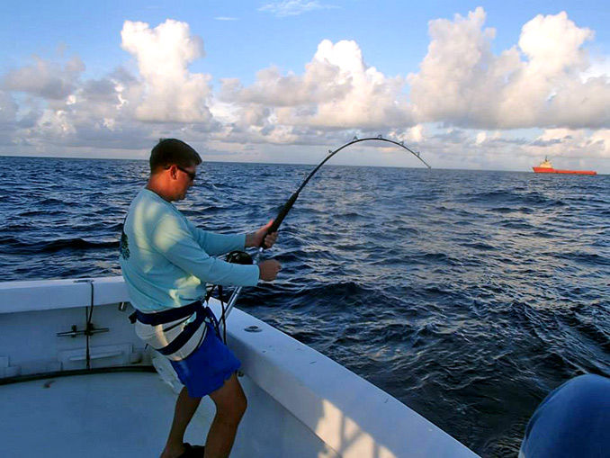 Man fishing on a boat in the ocean, wearing a blue shirt and shorts, with a bent fishing rod. Cargo ship visible on the horizon.