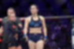 MMA fighter in blue UFC gear stands confidently in the octagon, with team members visible in the background. Arena atmosphere is focused.