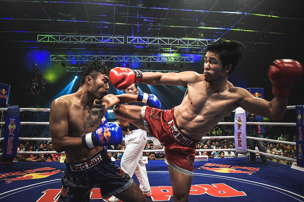 Two Muay Thai fighters compete in a ring. One delivers a high kick, wearing red gloves and shorts with "Red Bull" text. Intense atmosphere.