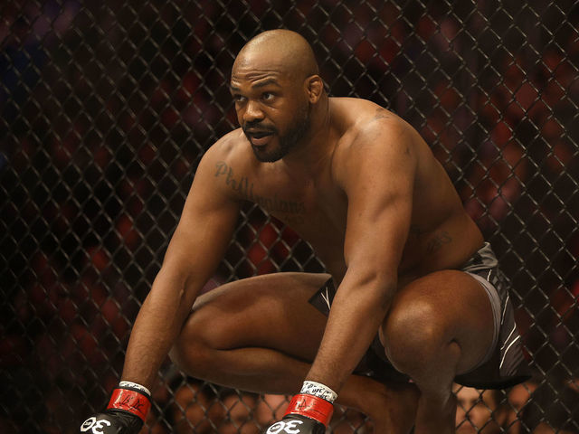 A man crouches in an MMA cage, wearing black trunks and red gloves. The background is a chain-link fence, and he's focused and ready.