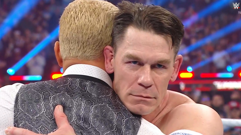 Two men embrace in a wrestling ring under bright lights. One has blond hair and a patterned suit. The mood is serious and intense.