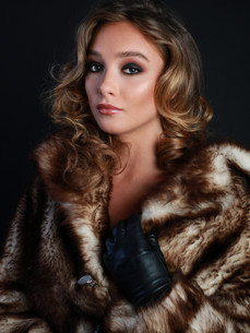 Woman with curly light brown hair wearing a fur jacket and one hand with a black glove on at the collar of the jacket.