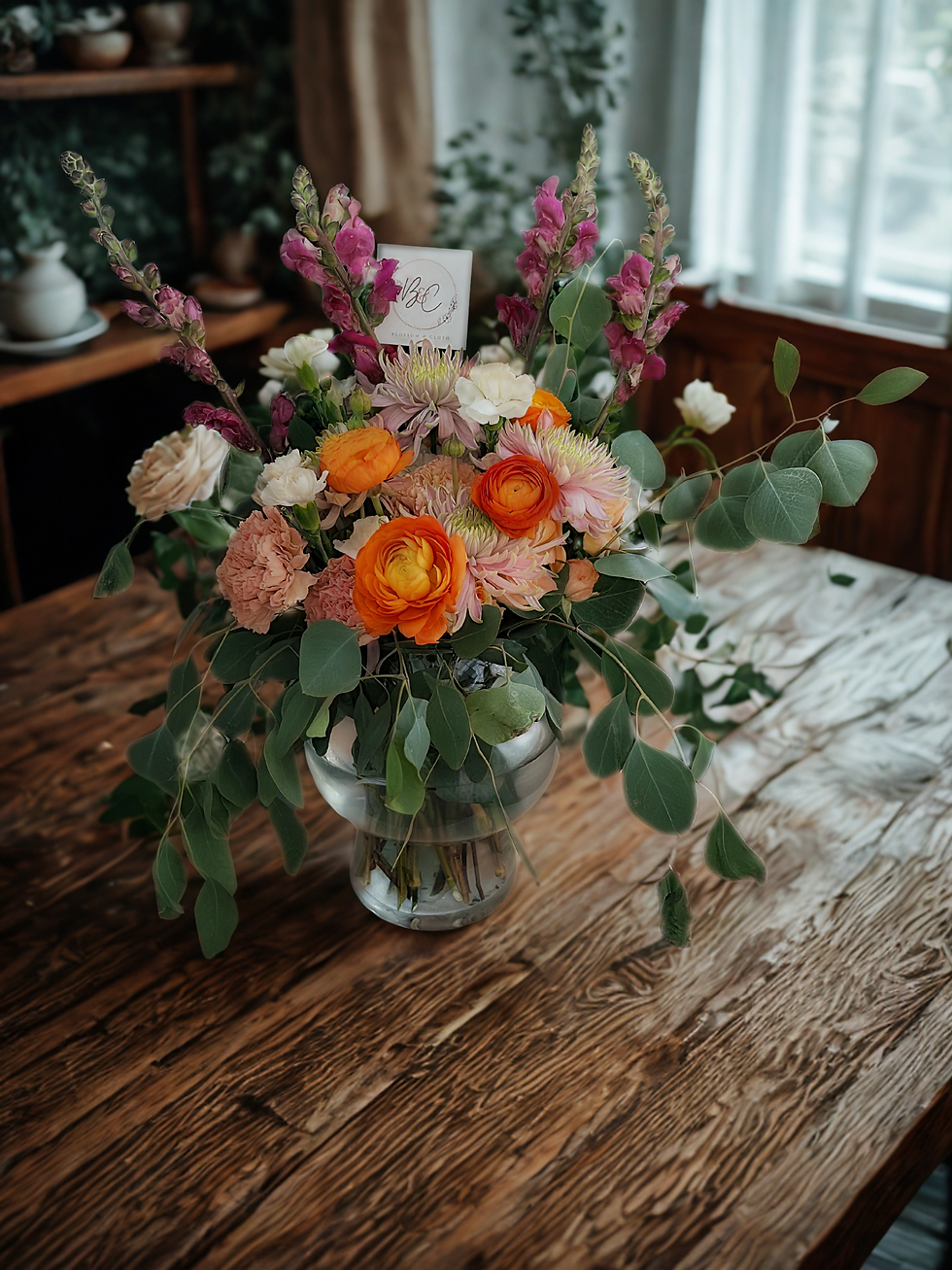 Colorful bouquet with orange, pink, and white flowers in a glass vase on a wooden table. Sunlight from a window in the background.