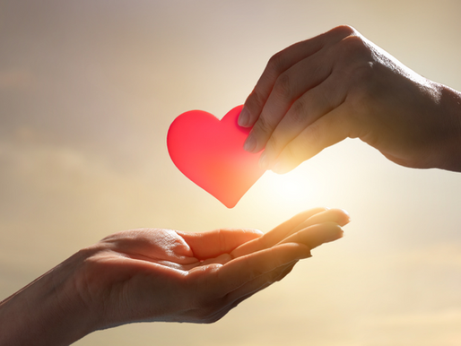 Two hands pass a red heart in warm light, symbolizing love and kindness against a soft, blurred background.
