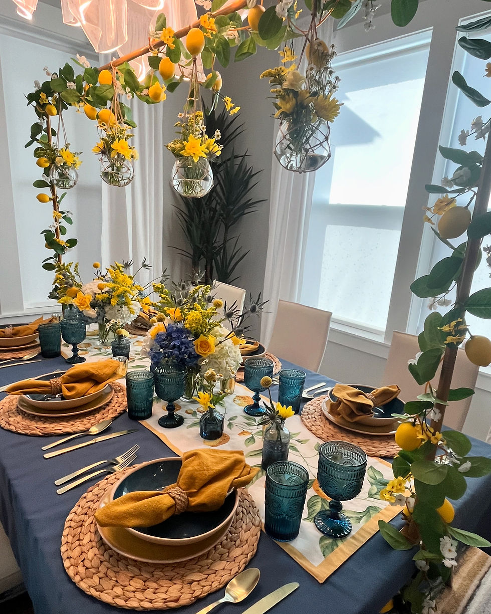 Elegant dining table with blue and yellow theme, floral centerpiece, hanging lemons, and yellow napkins. Light streams through a window.
