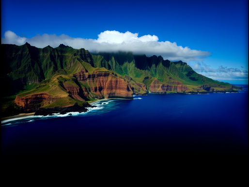 Aerial view of a lush green mountain range with cloud cover, next to a vibrant blue ocean. Coastal cliffs and waves create a serene scene.