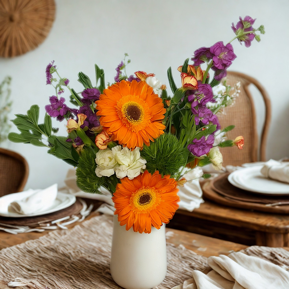 Vase of vibrant orange gerbera, purple and white flowers on a rustic dining table with woven placemats and white dishes, cozy setting.