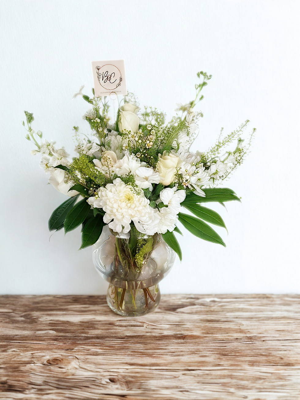 White flower bouquet in clear vase on wooden surface, with a beige card reading "V&F." Bright, minimalist background.