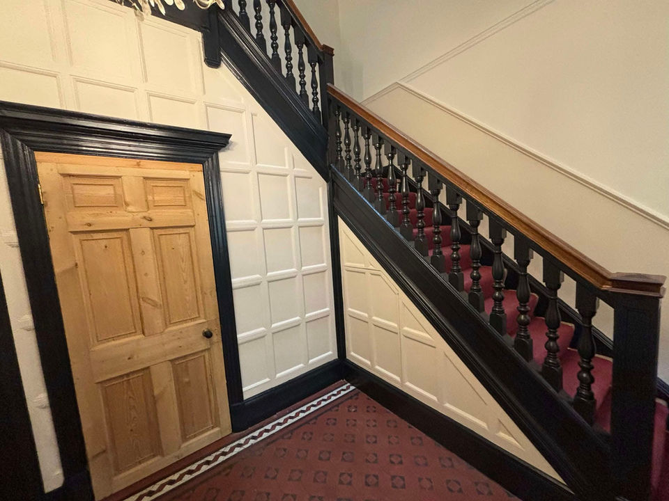 An older looking stairwell in a home, white walls with trim all in black, red carpet, wooden door