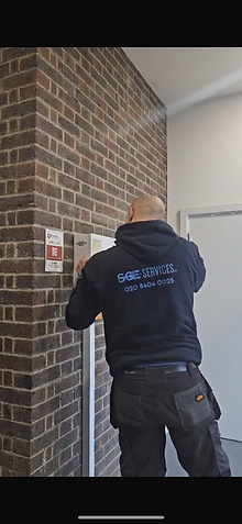 A person in a black hoodie with "SGE SERVICES" and a phone number is posting a paper on a brick wall indoors