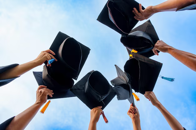 Wide photo of multiple hands holding graduation caps up toward a blue sky