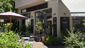 Aufnahme von einem Café und Kaufladen namens "Kebe". Der Laden ist umgeben von grünen Pflanzen und Sitzmöglichkeiten.