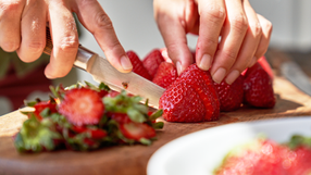 Nahaufnahme von zwei Händen, die mit einem Küchenmesser frische Erdbeeren aufschneiden