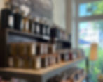 Regale im Laden von Kebe Living, sorgfältig bestückt mit handgemachten Feinkostprodukten wie Tapenade, Chutneys, Essigen und Gewürzen – dahinter ein großes Fenster mit Blick ins Grüne.