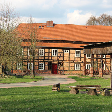 Blick über eine Wiese mit Feuerstelle auf ein zweistöckiges Fachwerkhaus aus hellbraunen Ziegeln. Daneben ein kahler Baum und Feldsteine
