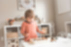 Young boy in orange shirt excitedly examines a wooden box with sand and shells in a bright room with white shelves and soft decor.