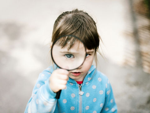 Child in a blue polka-dot jacket holds a magnifying glass, magnifying one eye. Neutral background, curious expression.