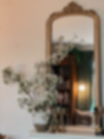Elegant mirror reflects a cozy room with a vintage lamp, books, and green curtains. A vase with white blossoms and brass candlesticks decorates a table.