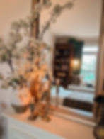 Vase with white blossoms and lit candles on a mantel, reflected in a mirror. Cozy room with a bookshelf, plaid blanket, and serene ambiance.