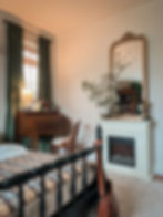 Cozy bedroom with a plaid-covered bed, elegant wooden desk, chair, ornate mirror with flowers, and a fireplace. Warm, inviting ambiance.