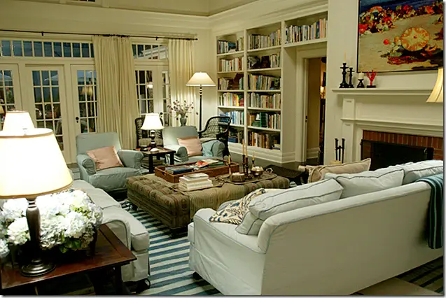 Cozy living room with blue sofas, striped rug, and bookshelves. Lamps illuminate the room. A colorful painting hangs above the fireplace.  Nancy Meyers inspired home from Something's Gotta Give, living room.