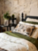A cozy bed with floral pillows and blanket, next to a table with a plant, vintage fan, and photo. The wall has paper sketches.