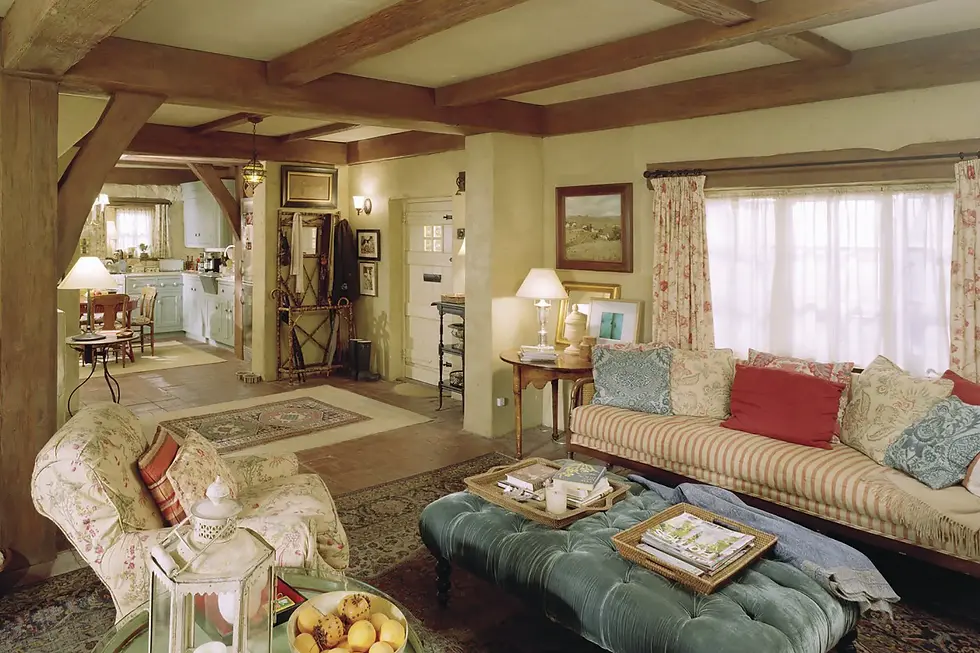 Cozy living room with floral patterns, striped sofa, and teal ottoman. Warm lighting, rustic wooden beams, and a bowl of oranges on a table.  The Holiday living room is a perfect example of Nancy Meyers style