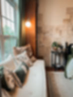 Cozy room with a daybed adorned with floral cushions, a vintage lamp, and a wall of sketches. Potted plant on a side table, natural light.