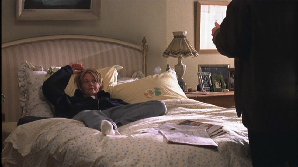The You've Got Mail bedroom has a fluffy, duvet covered bed with floral pillows. Framed photos and a lamp are on the bedside table.