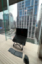 Ergonomic laptop setup on an adjustable stand with keyboard and mouse in front of a city skyline view through tall glass windows