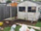 Before photo of small concrete block shed next to a vegetable garden dig site with a shovel.