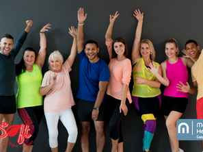 A diverse group of eight cheerful adults, dressed in colorful activewear, standing in front of a dark grey wall with their arms raised enthusiastically. The group includes people of different ages, genders, and ethnicities, all smiling and looking energetic. On the left bottom corner, the red QueSoft logo with the tagline "Comfort & Comfort" is displayed. On the right bottom corner, the McQuel Healthcare Continence Solutions logo and tagline "Comfort s Comfort" appear in a blue rounded rectangle.
