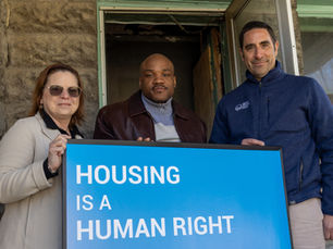 Cheryl Lawrence, Alderman Bobby Burns, and Rob Anthony stand in front of blue sign that reads "Housing is Human Right" on the steps of the Hovland Crt house.