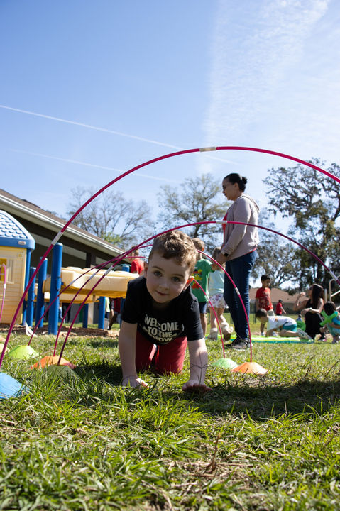 Boy crawls under a red arch, kids and a teacher, clear day, All Kids Academy.