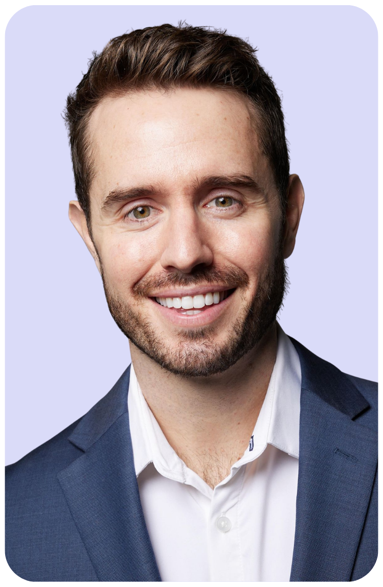 Smiling man in a navy suit and white shirt against a light lavender background, shown in a close-up portrait.