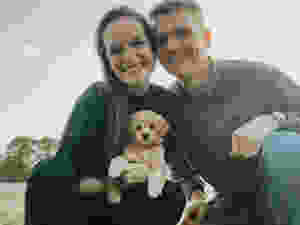 A family photo of a couple with their mini golden doodle puppy outside of a farm house.