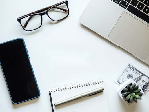 Valkoisella työpöydällä työvälineitä, kuten työpuhelin, tietokone, muistivihko ja silmälasit.