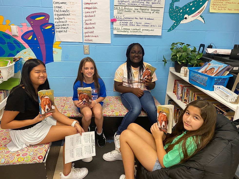 Four kids sit in a colorful classroom holding "Shiloh" books, with educational posters on the blue wall, creating a cheerful reading scene.