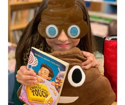Child wearing poop emoji glasses holds a book titled "The Chocolate Touch" and a matching pillow. Bookshelves in the blurred background.