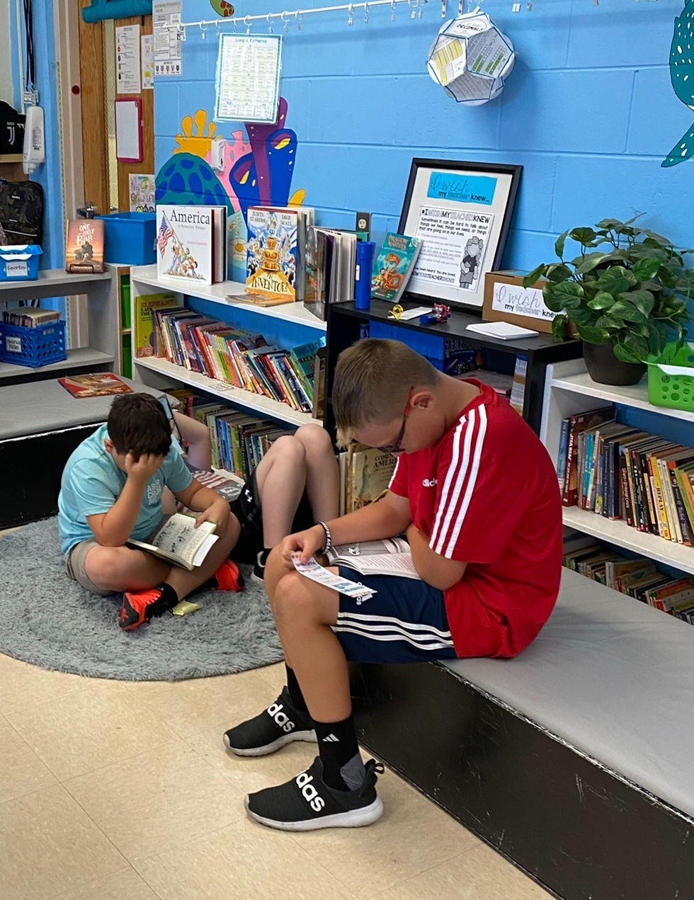 Two boys sitting and reading in a colorful library. One in a red shirt, the other in blue. Bookshelves and educational posters adorn the walls.